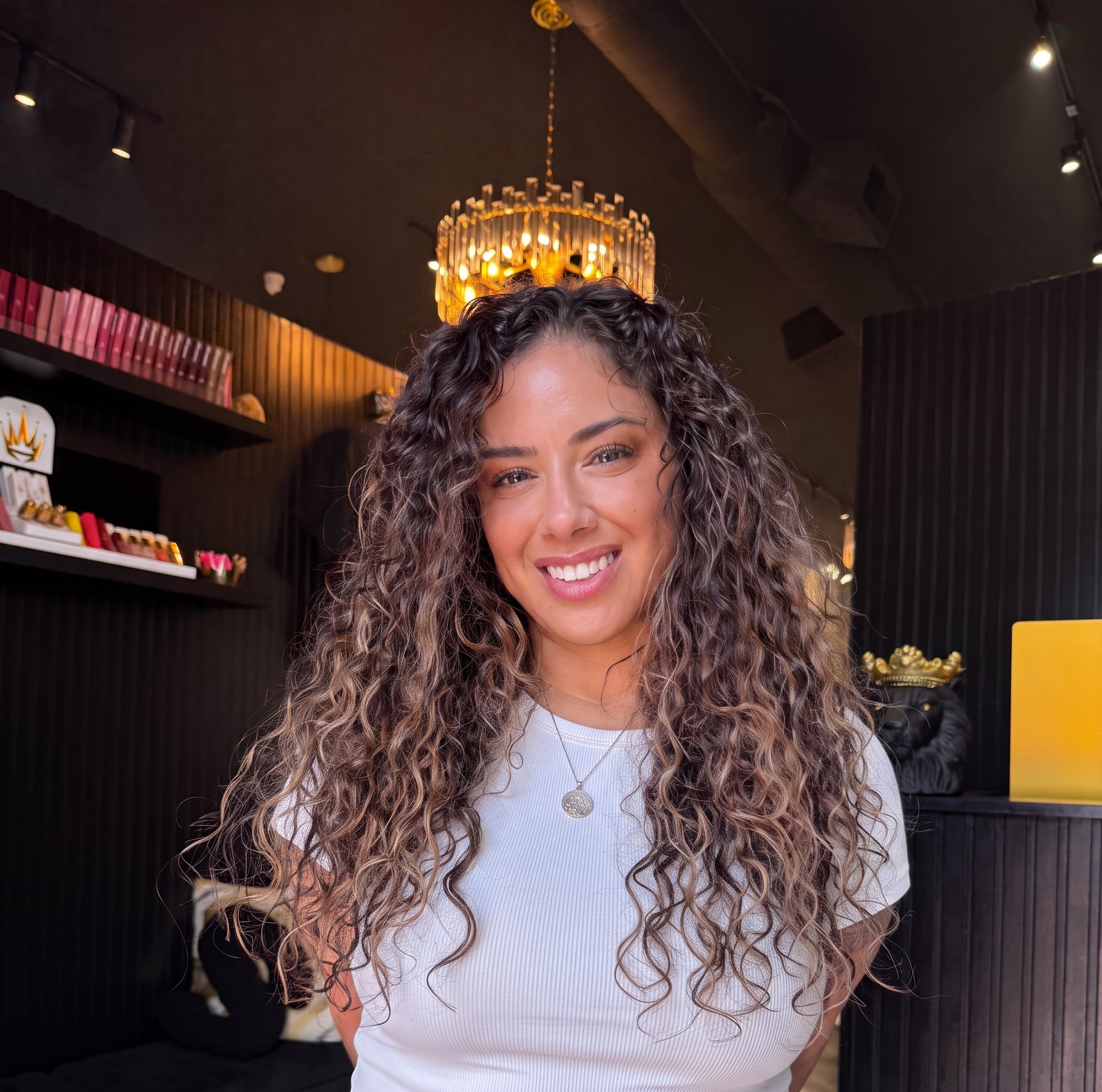 House of Hair client with long natural spiral curls in a white top beneath the salon's gold chandelier